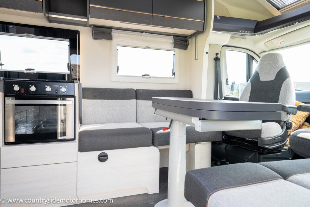 The image shows the interior of a 2021 Chausson 778 Premium motorhome with a modern design. There is a seating area with a gray upholstered bench, a dining table, and a swivel chair. A compact kitchen area features a built-in oven and cabinets. Large windows provide natural light.