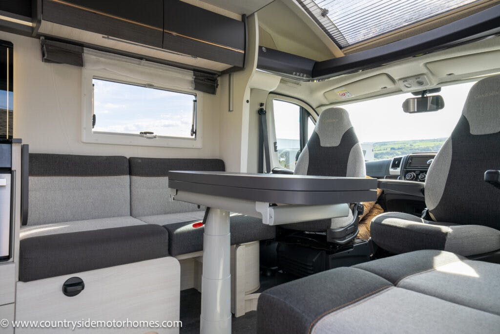The interior of the 2021 Chausson 778 Premium motorhome showcases a sophisticated gray and beige color scheme. Features include a table, L-shaped sofa with storage, driver's seat, and passenger seat. Large windows flood the space with natural light. The site logo appears in the bottom-left corner of the image.
