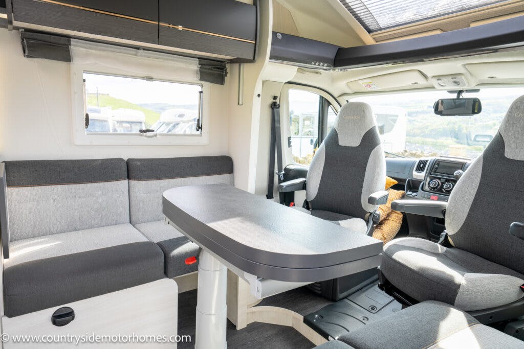 Interior of the 2021 Chausson 778 Premium motorhome featuring a seating area with gray and white upholstery, a foldable table, swiveling front seats, and overhead storage cabinets. The front windshield reveals a scenic view of the outdoors. The logo on the image reads www.countrysidemotorhomes.com.