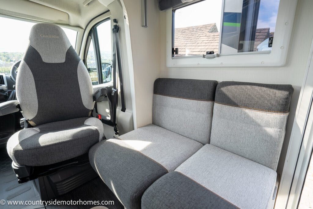 The image shows the interior of a 2021 Chausson 778 Premium motorhome featuring a single gray captain’s chair on the left and a two-seater gray and black fabric bench on the right. Sunlight is coming in through a window above the bench. A website URL is visible in the bottom-left corner.