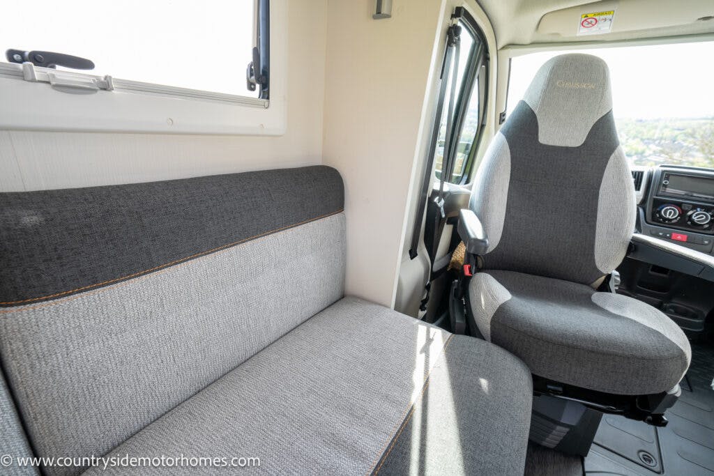 Interior of a 2021 Chausson 778 Premium motorhome featuring a gray upholstered bench seat and a driver's seat. The driver's seat is positioned next to the steering wheel and dashboard, while a window sits above the bench seat. This image showcases the refined interior from countryside motorhomes.