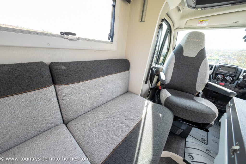 The interior of the 2021 Chausson 778 Premium motorhome features a grey sofa on the left and a driver’s seat on the right. A window above the sofa permits natural light, enhancing the bright ambiance created by wood flooring, with the dashboard and steering wheel visible in the background.