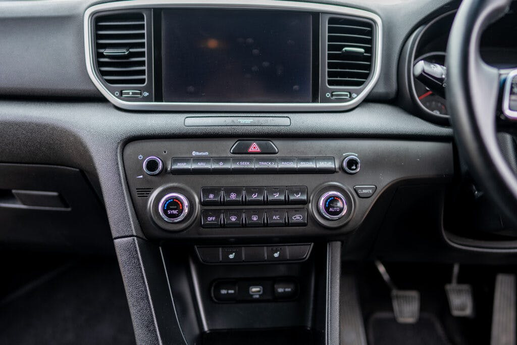 The 2021 Kia Sportage 1.6 GDi 2's dashboard features a central display screen, several buttons including a hazard light switch, climate control knobs, and a row of function buttons for media and air conditioning. Below are additional buttons and a slot, with the steering wheel and pedals partially visible.