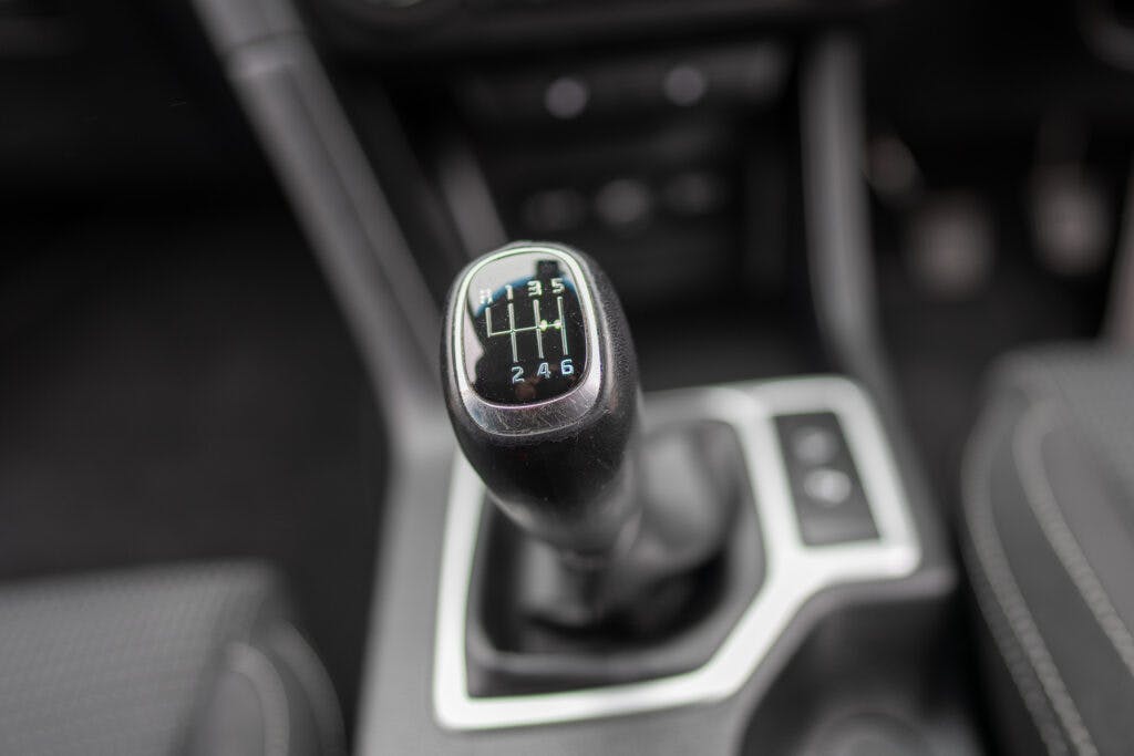 Close-up view of the manual gear shift knob in a 2021 Kia Sportage 1.6 GDi 2. The gear knob displays a 6-speed gear pattern, with reverse positioned on the left. The background reveals parts of the car's interior, including seats and control buttons.