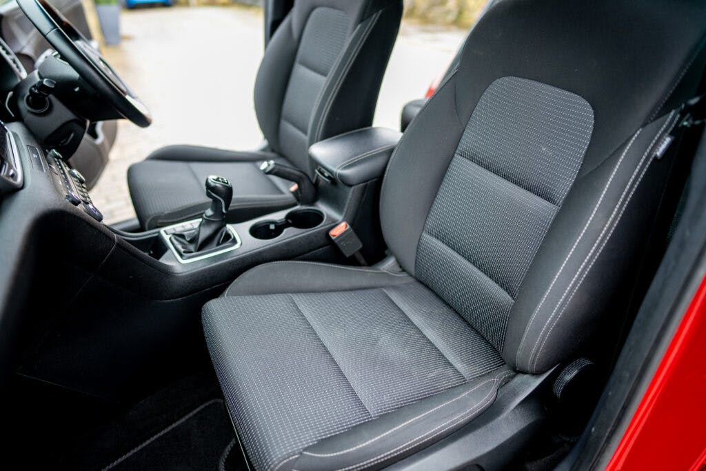 This image shows the interior of a 2021 Kia Sportage 1.6 GDi 2, focusing on the front seats and center console. The seats are upholstered in dark fabric with grey accents. The center console features two cup holders, the gear shift, and an armrest. The driver's seat has visible stitching.