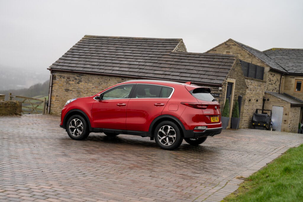 A red 2021 Kia Sportage 1.6 GDi 2 is parked on a wet brick driveway in front of a stone house with a tiled roof. The scene is overcast and the ground is wet, suggesting recent rain. The house has a few windows and a double garage. The vehicle's license plate is visible.