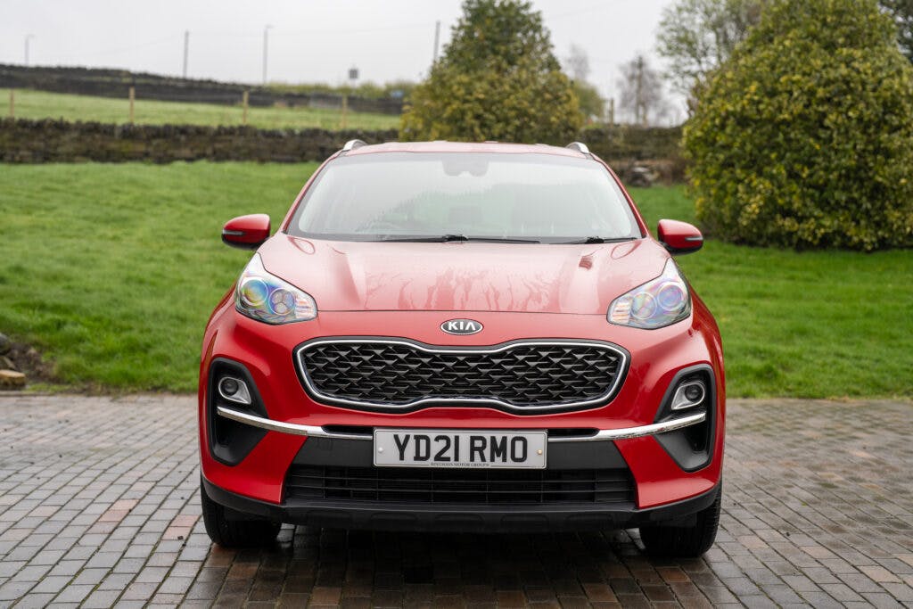 A red 2021 Kia Sportage 1.6 GDi 2 is parked on a brick driveway. The vehicle's license plate reads "YD21 RMO." The background includes greenery, a stone wall, and a slightly overcast sky.
