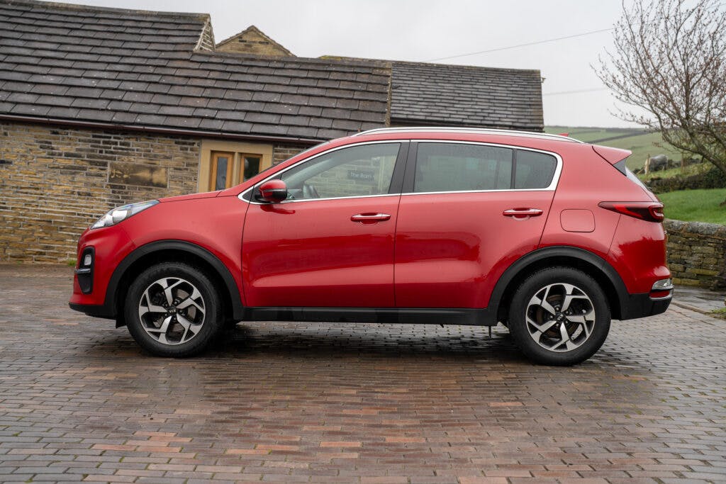 A 2021 Kia Sportage 1.6 GDi 2 is parked on a cobblestone driveway in front of a stone house with a tiled roof. The red SUV, viewed from the side, showcases its four doors and tinted windows. Leafless trees and a grassy area are visible in the background.