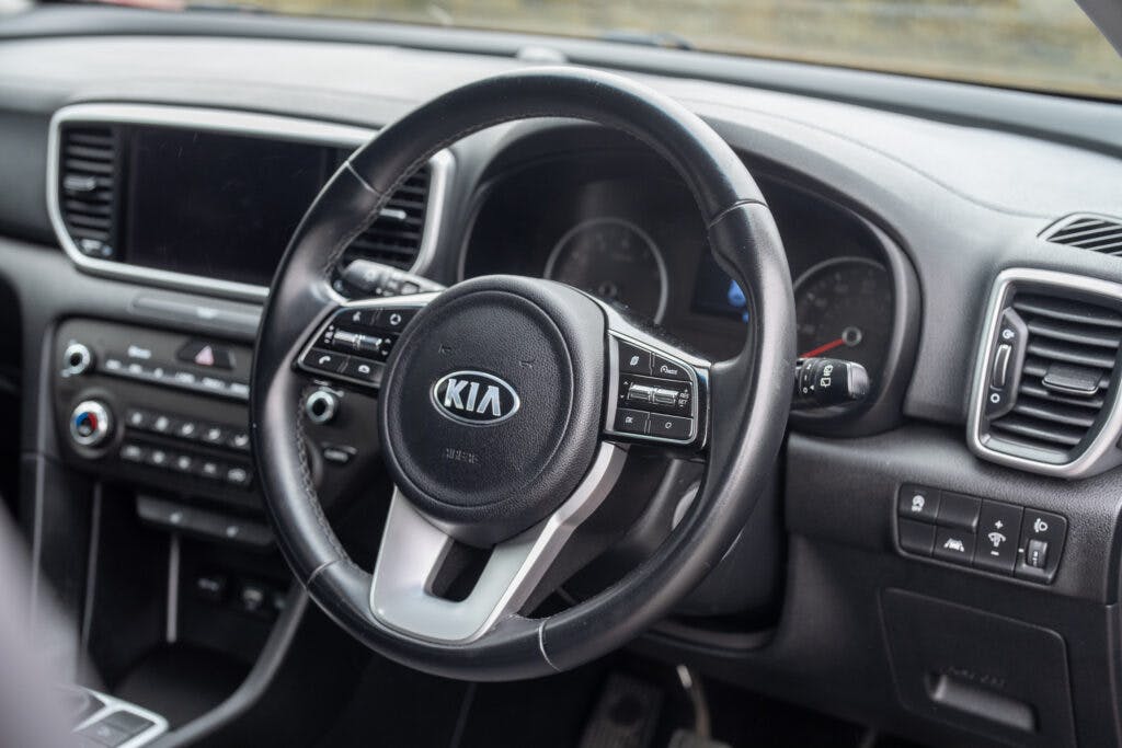Interior view of a 2021 Kia Sportage 1.6 GDi 2 showing the steering wheel, control buttons, a digital display, and various dashboard components including air vents, buttons, and knobs. The steering wheel has the Kia logo at the center.