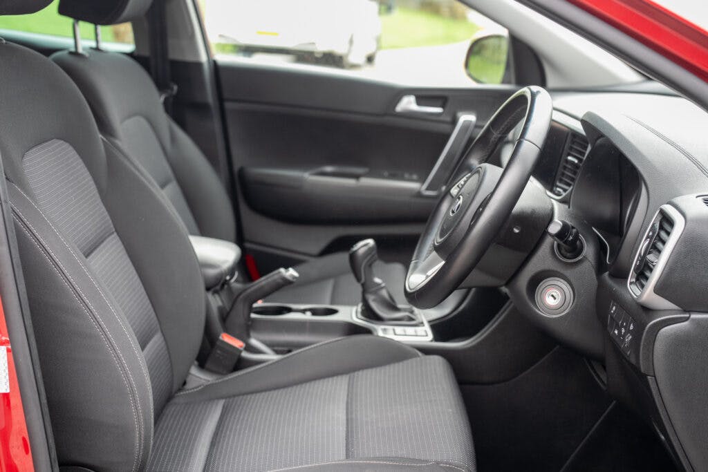 This image shows the interior of a 2021 Kia Sportage 1.6 GDi 2 from the driver's side perspective. It includes a steering wheel, dashboard, gear shift, and two front seats in dark gray fabric. The car is parked with the driver’s side door open, revealing part of the exterior.