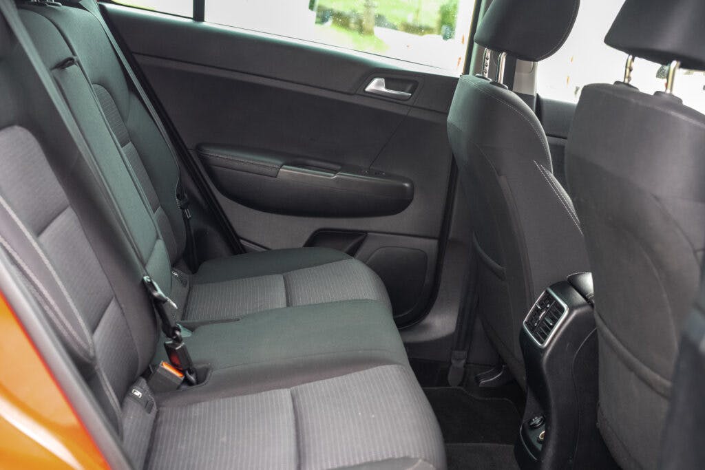 The image shows the interior of a 2021 Kia Sportage 1.6 GDi 2 viewed from the back seat. The seats are covered in dark fabric upholstery. The door has a silver handle, and there are air vents at the back of the center console. A window shows greenery outside.
