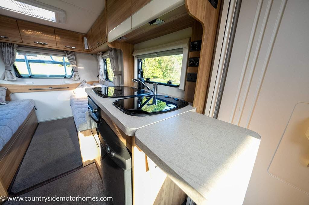 The image shows the interior of a 2015 Elddis Accordo 140 motorhome, specifically the kitchen and lounge area. The kitchen features a countertop with a sink and stove, cabinets, and appliances. The lounge area includes comfortable seating with cushions and a window providing natural light.