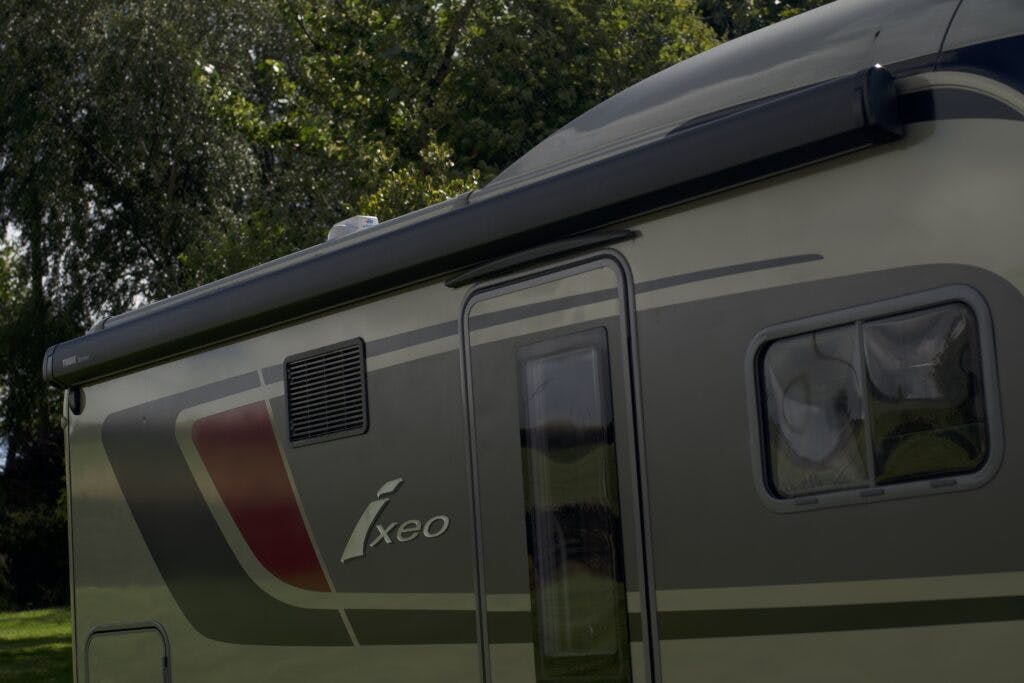 The image shows the side of a gray 2018 Burstner Ixeo TL680 G camper van parked outdoors. The camper features a dark awning, reflective windows, and a partial view of a ventilation panel. Trees are visible in the background under a natural daylight setting.