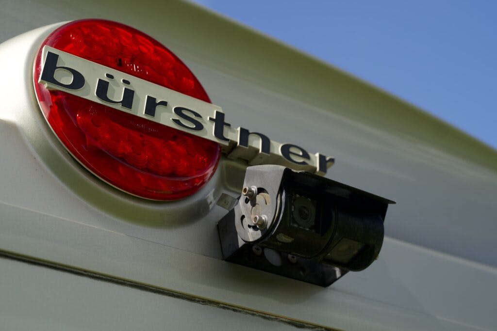 Close-up of the back of a 2018 Burstner Ixeo TL680 G, featuring a red light and a rear-view camera. The word "Bürstner" is prominently displayed above the camera in metallic letters. The background shows a clear blue sky.