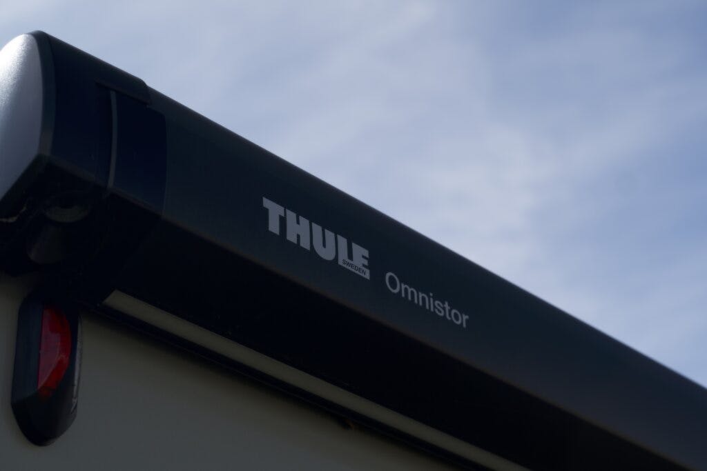 Close-up of a Thule Omnistor awning on the 2018 Burstner Ixeo TL680 G. The frame is mostly black with white lettering indicating the brand name "Thule" and model "Omnistor." The sky in the background is clear and blue.