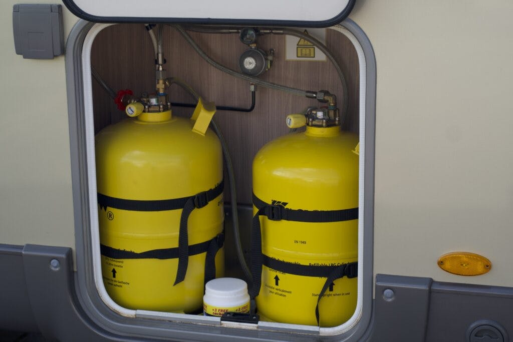 The image shows the open compartment of a 2018 Burstner Ixeo TL680 G containing two large yellow propane tanks secured with black straps. There is also a small white container placed at the bottom of the compartment. Various hoses and connectors are visible.