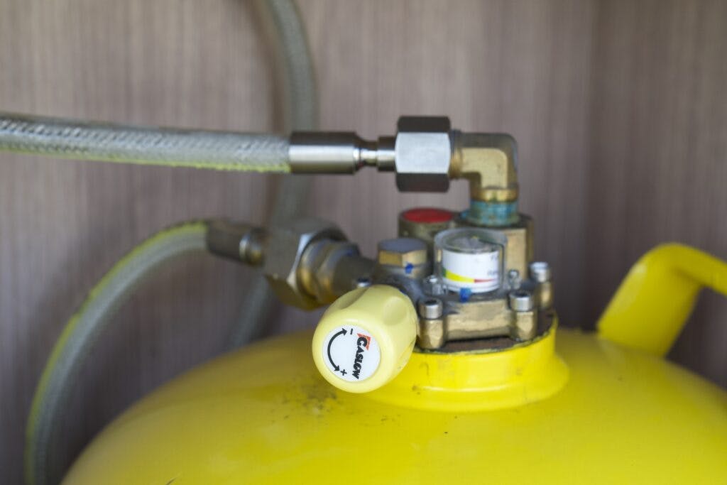 Close-up view of a yellow gas cylinder valve assembly, reminiscent of the precision found in a 2018 Burstner Ixeo TL680 G, showing various connections and fittings including a pressure gauge and safety valve. The hose attached to the valve is transparent with visible reinforcement against a wooden surface background.
