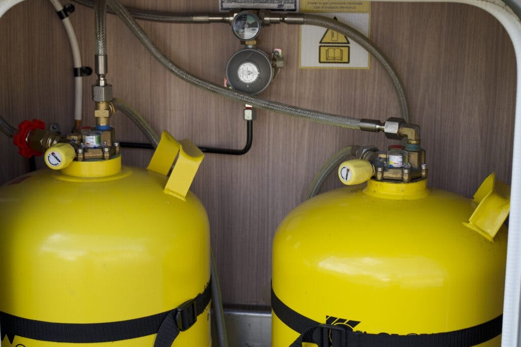 The image depicts two large yellow gas cylinders, reminiscent of those in a 2018 Burstner Ixeo TL680 G, connected by metal hoses and set against a wooden backdrop. Each cylinder features a gauge on top and is secured with black straps. A warning sign is visible in the background.