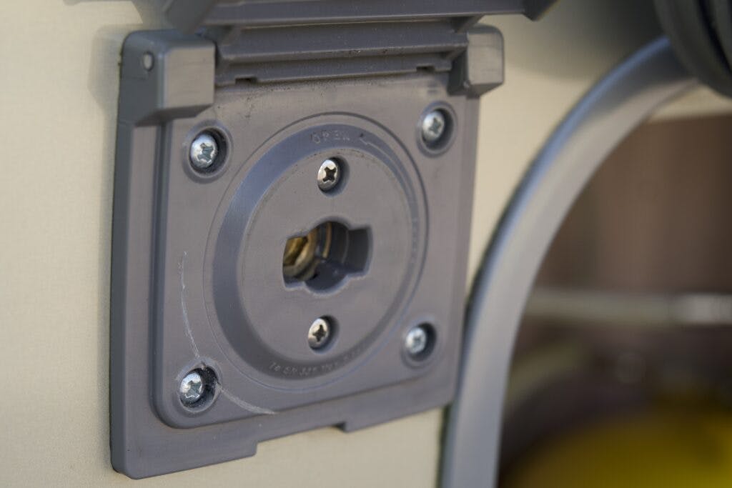 A close-up photo of an outdoor electrical outlet with a protective cover on a 2018 Burstner Ixeo TL680 G. The outlet has three screw holes and an open slot in the center. The cover is flipped up, and there is a wire running to the right side of the outlet.