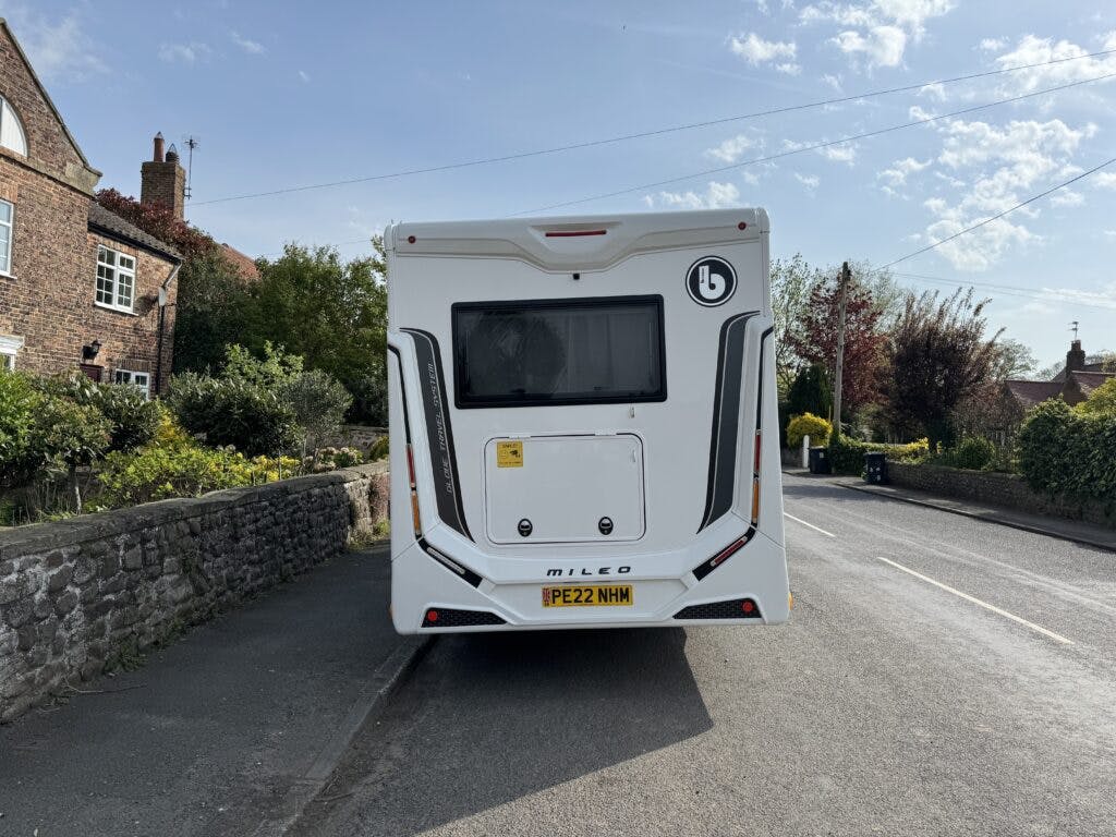 Rear view of a white 2022 Benimar Mileo 282 motorhome with the license plate "PE22 NHM" parked on a residential street. The motorhome has the brand logo and model name "Mileo" visible, with brick houses and stone walls lining the street under a partly cloudy sky.