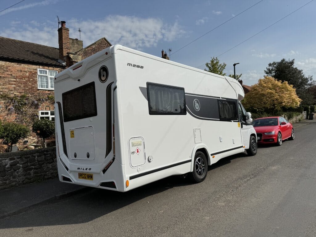 A white 2022 Benimar Mileo 282 motorhome with license plate "PZ22 YWM" is parked on a residential street. There is a house made of brick and a red car in the background. The sky is mostly clear with a few clouds.
