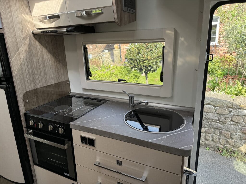 This image shows the modern kitchen area of a 2022 Benimar Mileo 282 RV featuring a stovetop, oven, and sink with a metal faucet. There is a window above the sink that provides a view of the outdoors, including a stone wall and greenery. The surroundings appear clean and organized.