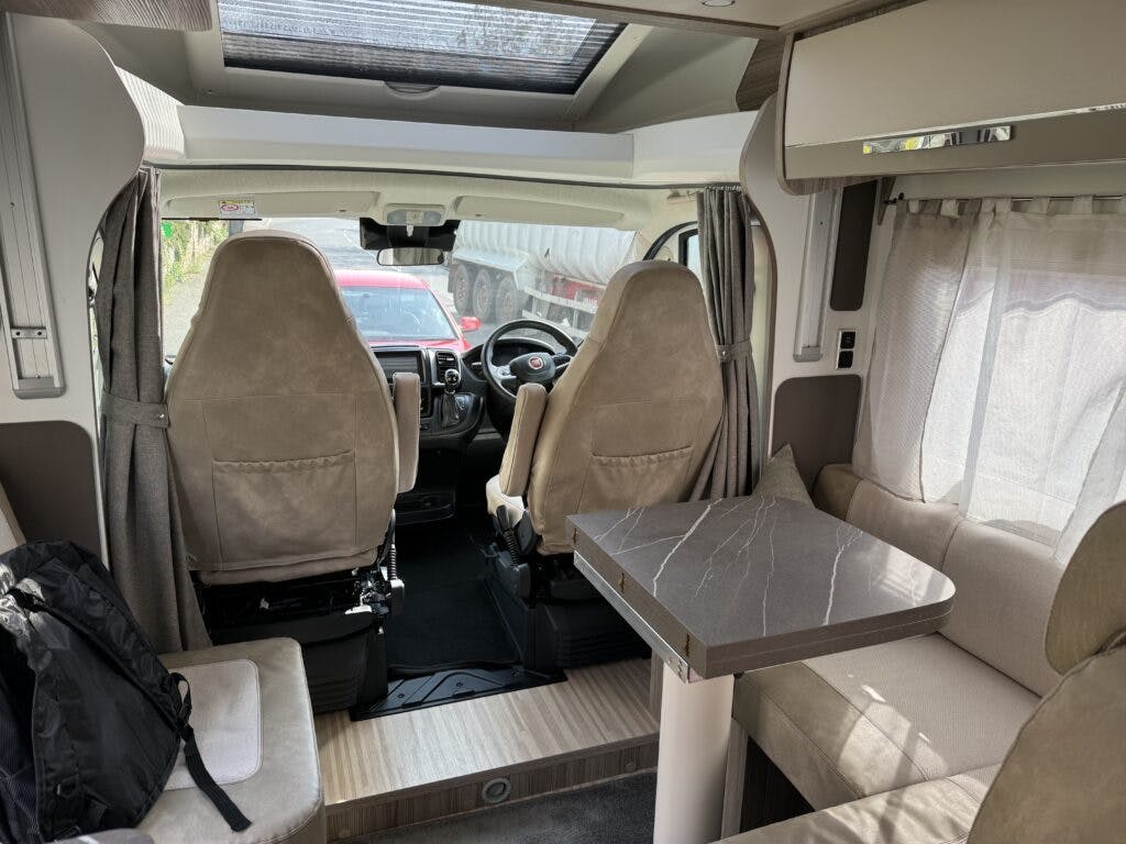 The interior of the 2022 Benimar Mileo 282 motorhome features two front seats, a dashboard with a steering wheel, and a small table with surrounding seating in the middle. A backpack is placed on one of the seats. The view outside shows a parked truck.