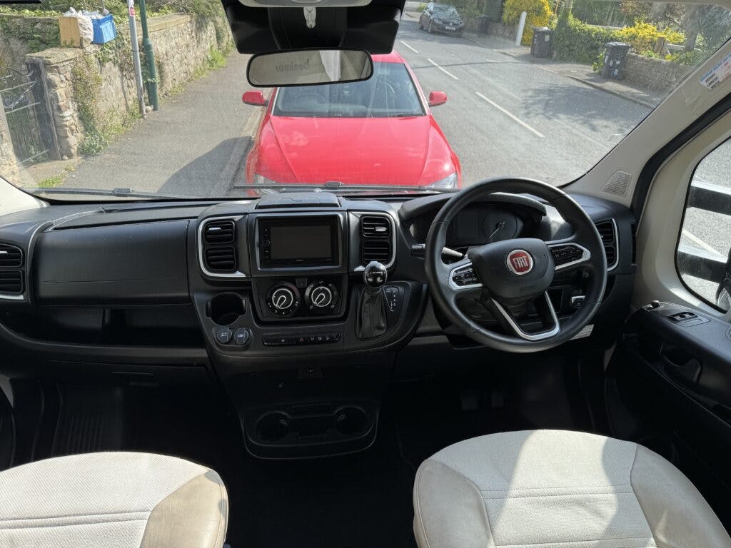 Interior view of the 2022 Benimar Mileo 282's driver area, showcasing a steering wheel with the Fiat logo, a dashboard with a central digital display, various controls and air vents. The front window shows a red car parked ahead on a residential street.