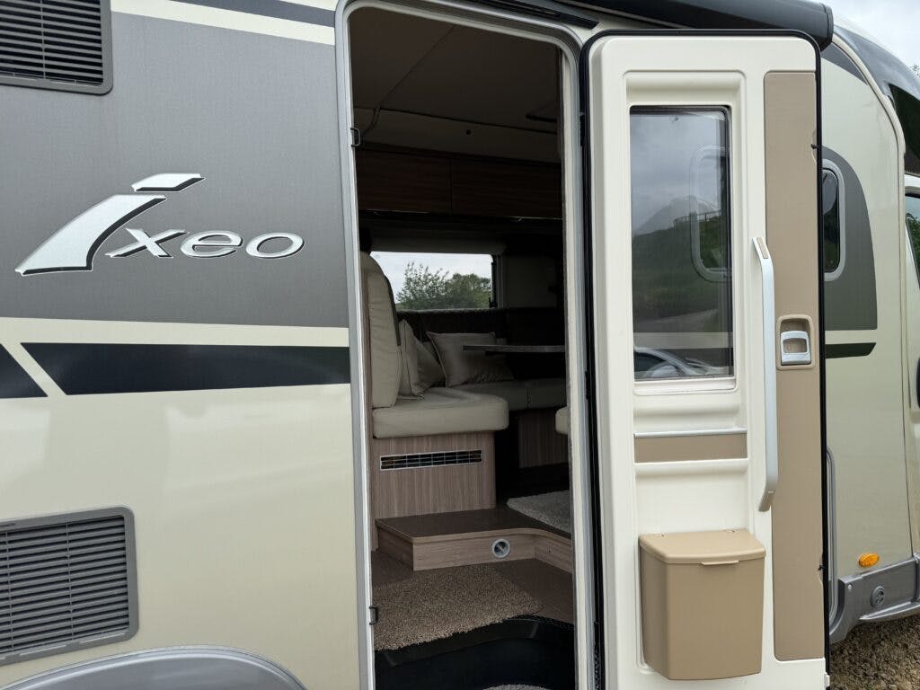 A close-up view of an open beige and gray camper van door with "Ixeo" branding. The interior of the 2018 Burstner Ixeo TL680 G reveals cushioned seating, a small dining area, and a step. A transparent window and a brown storage box are attached to the door.