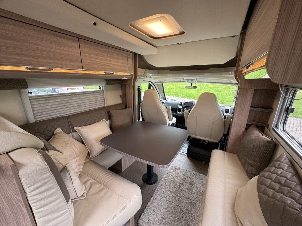 Interior view of the 2018 Burstner Ixeo TL680 G camper van featuring beige and brown tones. It includes a seating area with cushioned benches, a table in the center, two front seats, and overhead storage cabinets. There is a window on the left side with a scenic grass view outside.