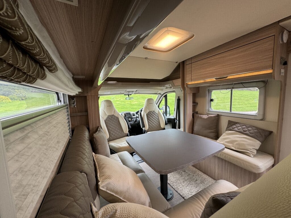 The image shows the interior of a 2018 Burstner Ixeo TL680 G camper van, featuring a dining area with cushioned seating, a rectangular table, and a window with blinds. The seats are upholstered in a beige and light brown color scheme. The front cab is visible in the background.