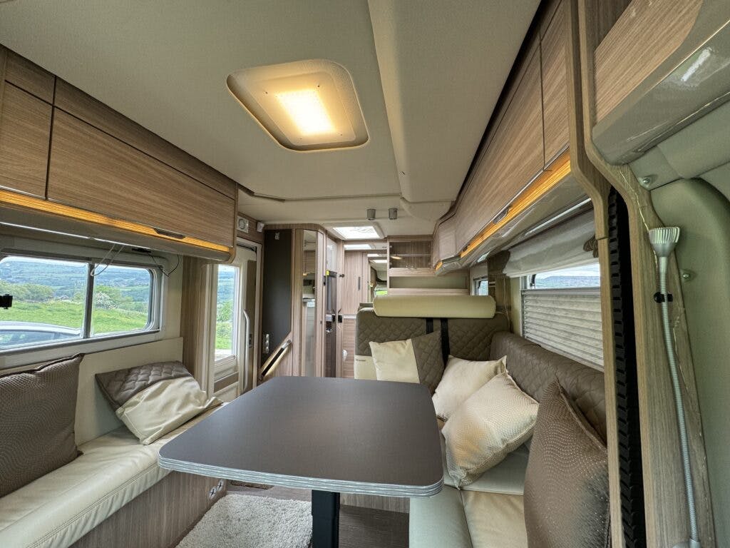 Interior of a 2018 Burstner Ixeo TL680 G RV featuring a dining area with a black table and cushioned beige seating. Overhead cabinets line the walls, and large windows provide views of greenery outside. A ceiling light and a doorway leading to another area are visible.