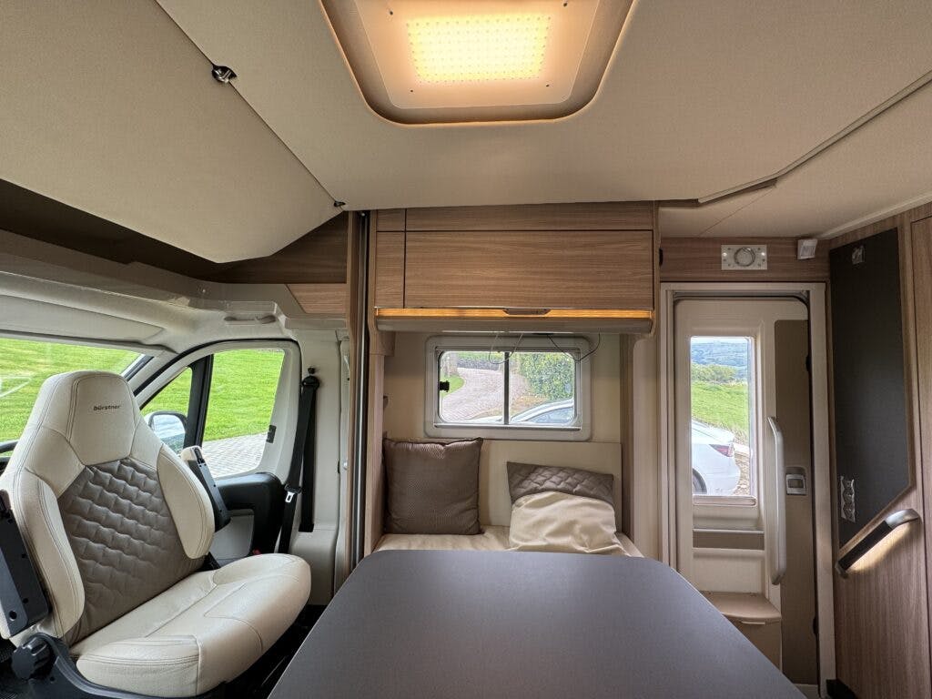 The interior of the 2018 Burstner Ixeo TL680 G camper van features a rotating driver's seat upholstered in white with a diamond pattern, a table, overhead storage cabinets, and a window with cushions below that bathe the space in natural light. The van door is partially visible on the right.