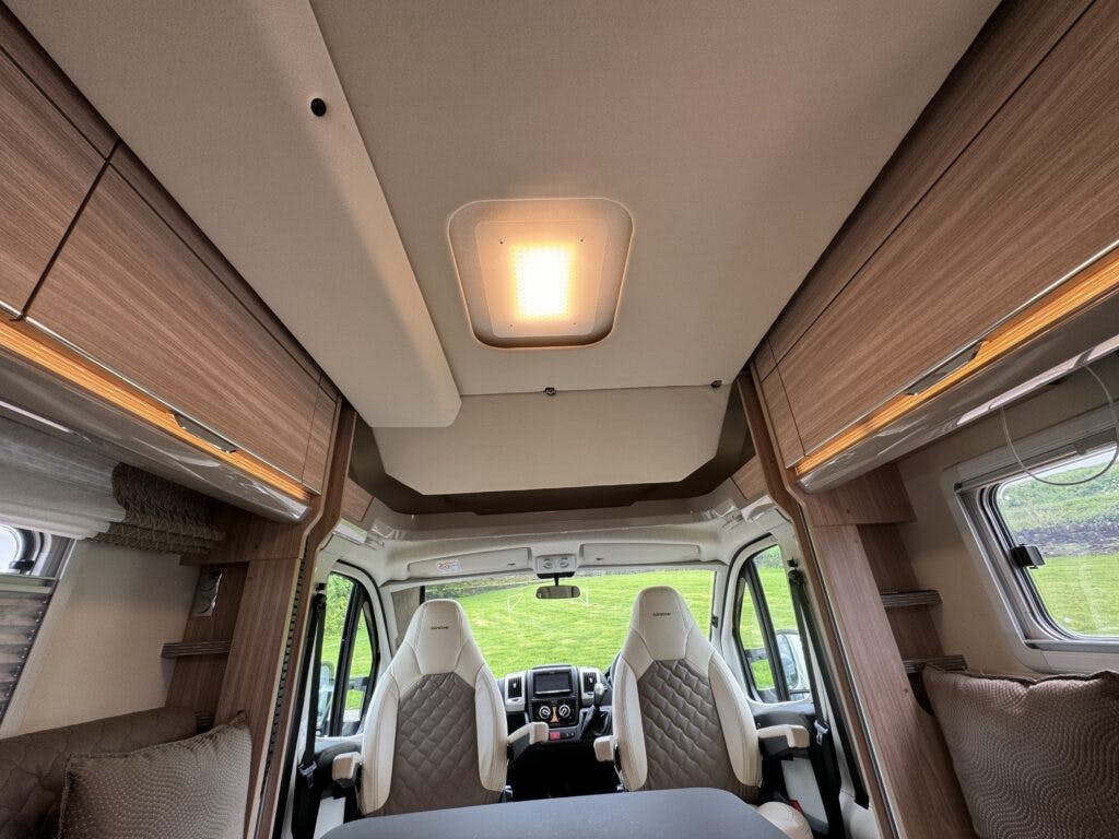 Interior view of the 2018 Burstner Ixeo TL680 G motorhome showcasing the ceiling, a central light fixture, two front seats with diamond-patterned upholstery, and surrounding wood-finished storage cabinets. A window with partial blinds is visible on the right side.