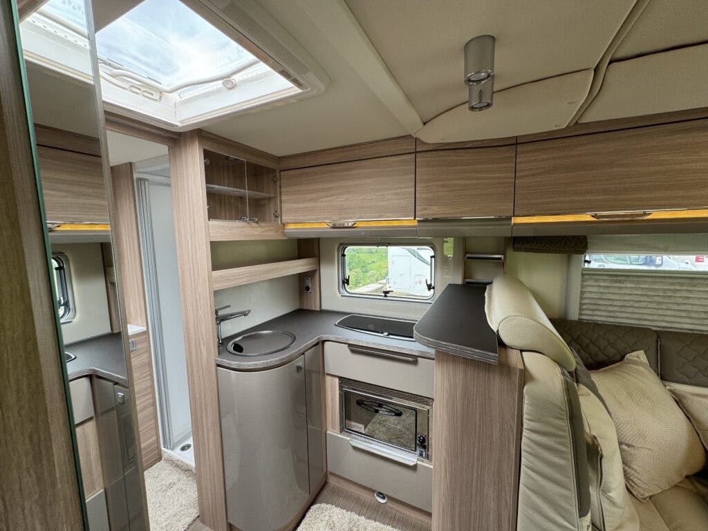A compact RV kitchen in the 2018 Burstner Ixeo TL680 G features wood-finished cabinets, a small sink, a microwave, and a refrigerator. A skylight is visible overhead, and a window offers a view outside. To the right, there is a cushioned seating area.