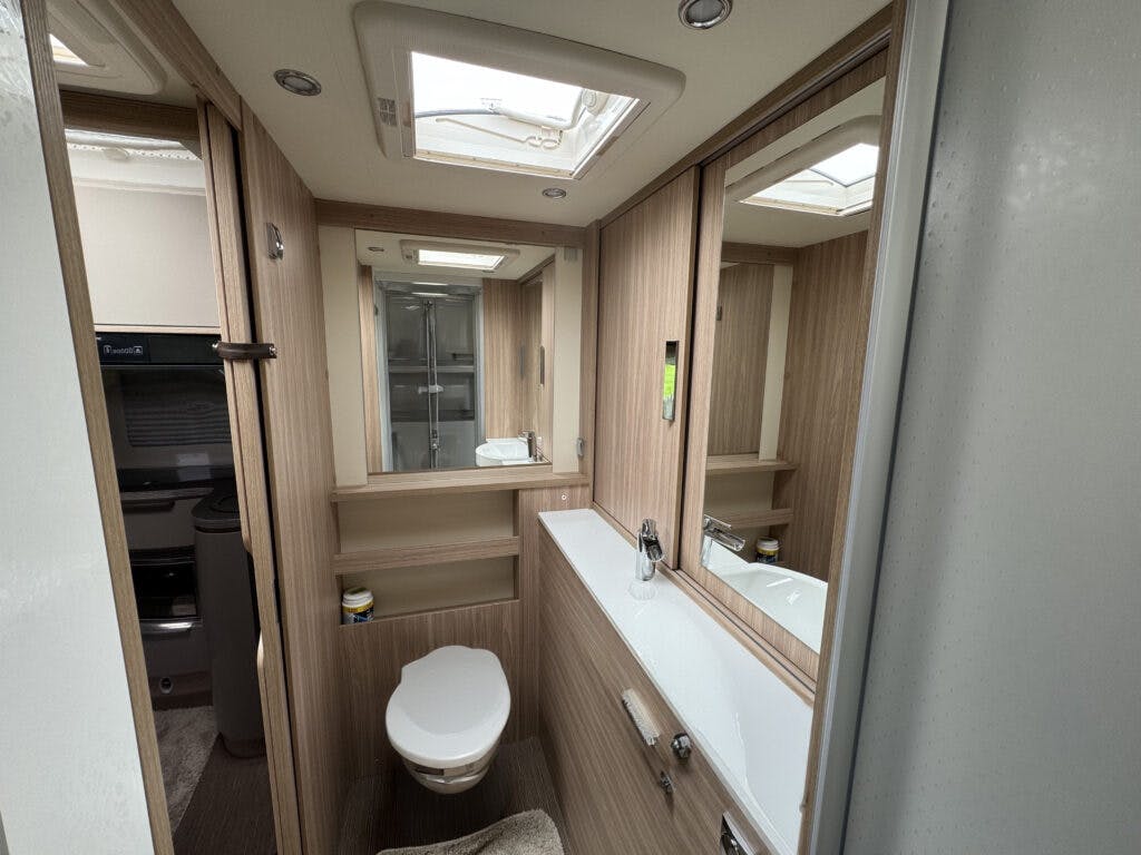 A modern RV bathroom in the 2018 Burstner Ixeo TL680 G features wood-panelled walls. The room includes a toilet, a sink with a faucet and a mirror above it, and a shower area enclosed by glass doors. Overhead lighting and natural light from a skylight illuminate the space.