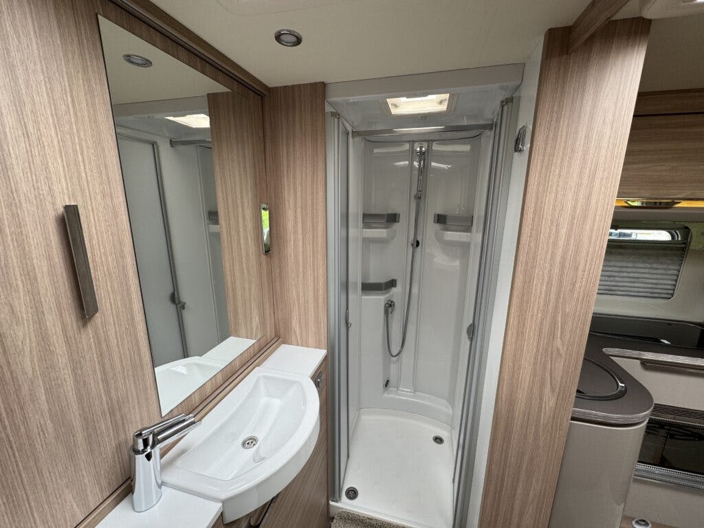 The 2018 Burstner Ixeo TL680 G boasts a modern bathroom with a shower featuring sliding doors, built-in shelves, and a handheld showerhead. A small sink with a mirror above it and stylish wood paneling complete the look. Adjacent to the bathroom is part of the living area with cabinets and a window.