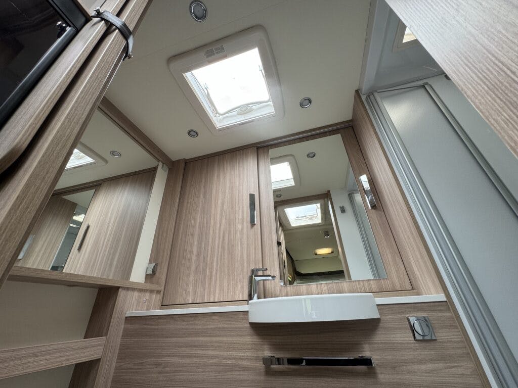 Interior of a modern 2018 Burstner Ixeo TL680 G RV bathroom with wooden cabinets, a rectangular sink, and a mirrored cabinet above. A small skylight and recessed lighting provide illumination. A sliding door to the shower is visible on the right side.