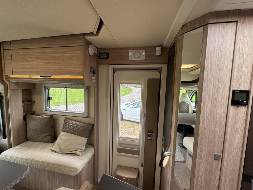 Inside view of a compact living area in the 2018 Burstner Ixeo TL680 G camper van. On the left, a cushioned bench with pillows sits below a storage cabinet. In the center, there's a door leading outside. To the right, you can see a partial view of the bathroom entrance.