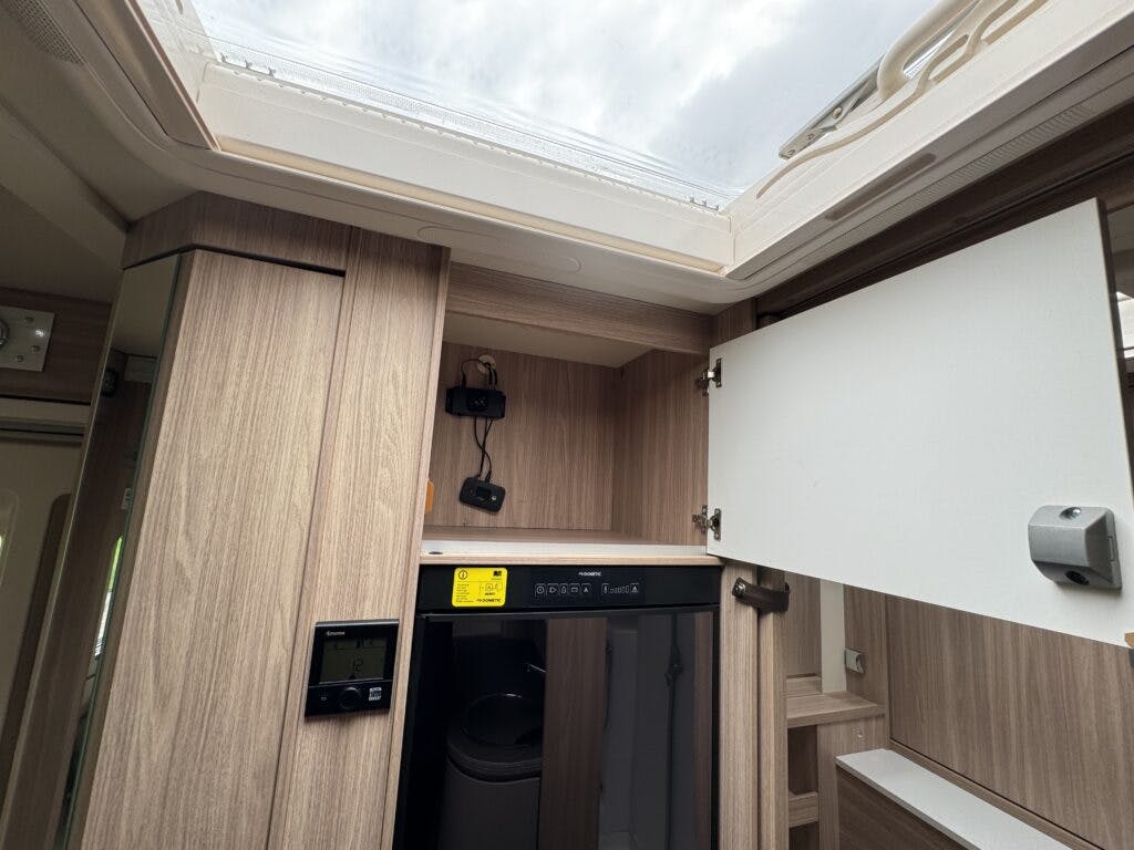 The image shows the interior of a modern 2018 Burstner Ixeo TL680 G RV. There is a small refrigerator with a digital display above it and an open cabinet door revealing a storage area. The interior features a light wood finish, and there is a large skylight on the ceiling.