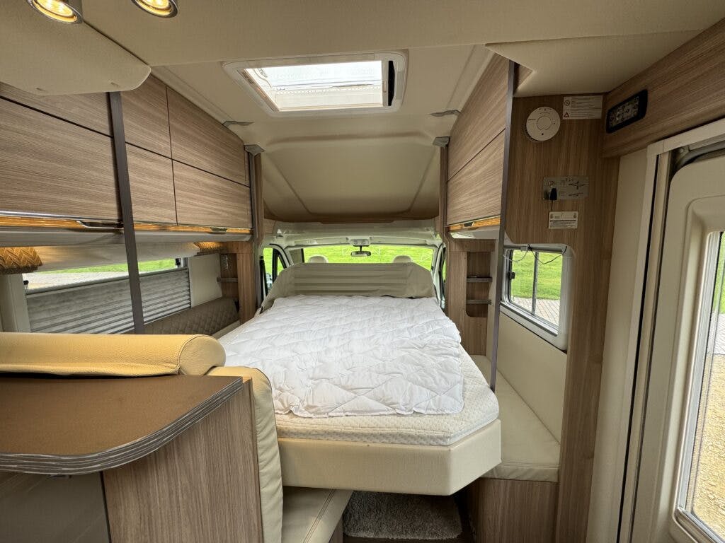 Interior of the 2018 Burstner Ixeo TL680 G camper van featuring a neatly made bed with a white comforter. The space includes wooden cabinetry above and beside the bed, a window with a rolled-up shade on the side, and a skylight providing natural light.