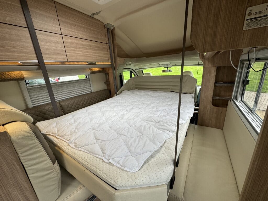 Interior view of the 2018 Burstner Ixeo TL680 G camper van featuring a neatly made bed with white bedding. The space includes overhead storage cabinets, light-colored upholstery, and windows on the side, offering a cozy and functional living area within the vehicle.