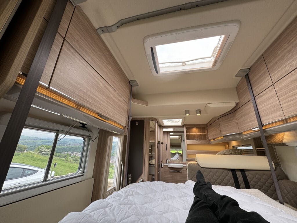 The image shows the interior of a 2018 Burstner Ixeo TL680 G camper van from the perspective of someone lying on the bed. The ceiling features a skylight, and there are overhead storage cabinets. A window to the left reveals a scenic view outside. The person's legs are visible in the foreground.