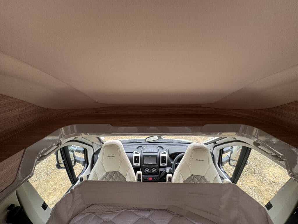 Interior view of the 2018 Burstner Ixeo TL680 G camper van from the rear, showcasing the driver and passenger seats with white covers. The dashboard and steering wheel are visible. A raised bed with a white blanket is above the cabin area, and the van's door is open, revealing a rocky ground outside.