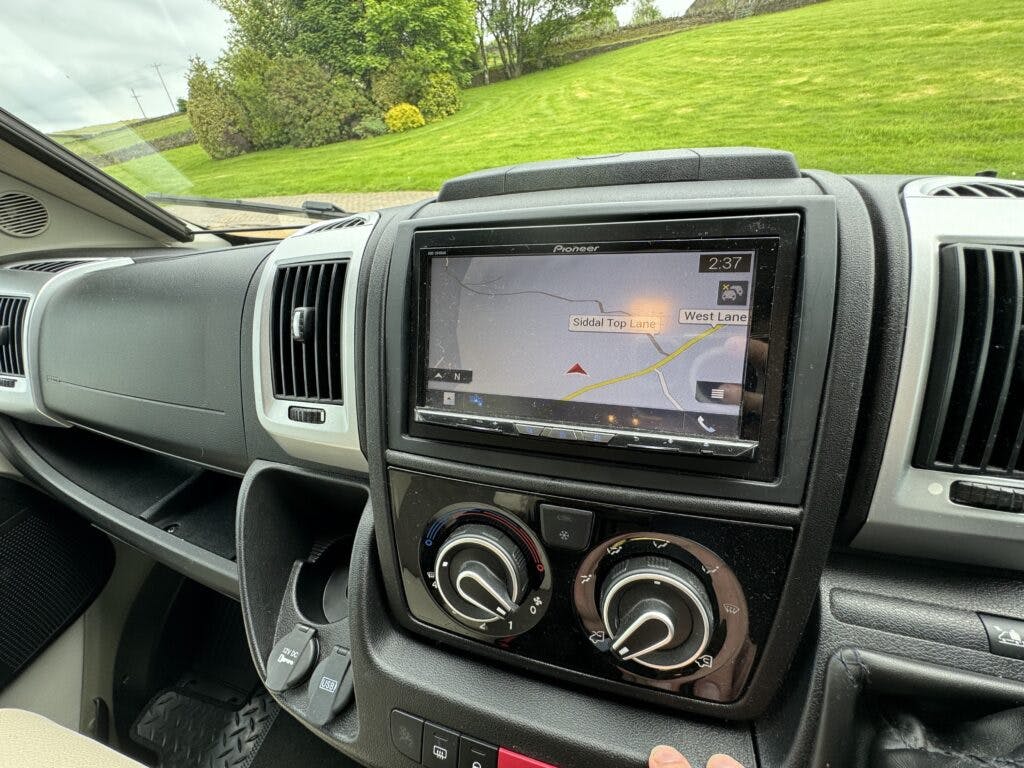 A car dashboard from the 2018 Burstner Ixeo TL680 G features a GPS navigation system displaying a route with street names "Siddal Top Lane" and "West Lane." The dashboard includes climate control dials, and the display time reads 2:37, all set against a backdrop of grassy surroundings.