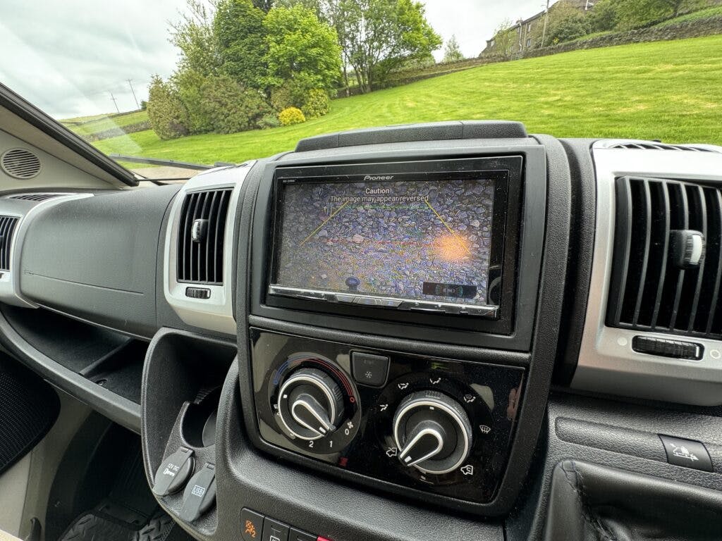 A car dashboard in the 2018 Burstner Ixeo TL680 G features an infotainment system displaying a rearview camera's footage. The screen shows reverse guidelines, and below it are two control knobs for climate control settings. The surrounding environment visible through the windows includes green grass and trees.