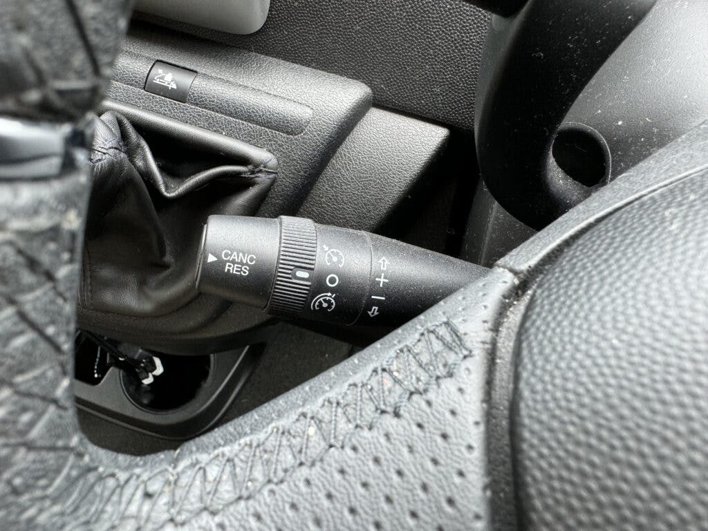 Close-up of a car's cruise control lever on the steering column in a 2018 Burstner Ixeo TL680 G. The lever displays symbols and text such as "CANC," "RES," and several control icons. The surrounding area includes part of the steering wheel and leather gear shift cover.