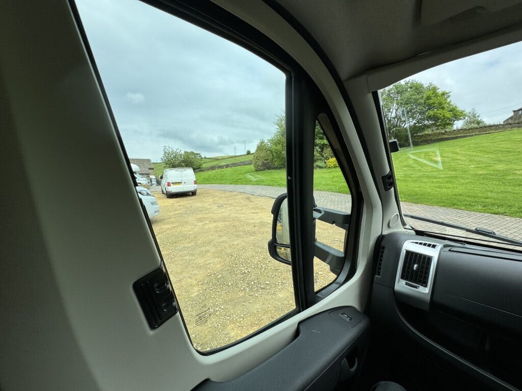 View from inside a 2018 Burstner Ixeo TL680 G looking out through the passenger side window. Outside, there are parked vehicles on a dirt path and well-maintained grassy areas. Trees and cloudy skies are visible in the background.