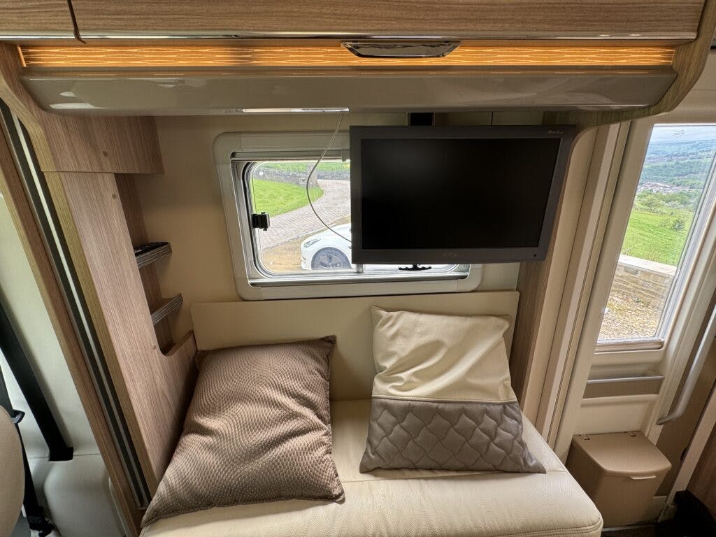 The image shows the interior of a 2018 Burstner Ixeo TL680 G motorhome with a small couch, two pillows, a mounted flat-screen TV, and a window with a scenic view. Light wooden cabinets and ambient lighting are visible above the TV.