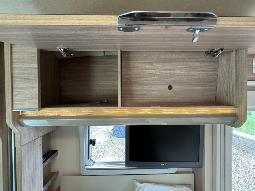 Inside the 2018 Burstner Ixeo TL680 G, a wooden overhead storage cabinet with a lifted door graces the recreational vehicle. Below the cabinet, a flat-screen television is mounted on the wall next to a small window. The interior is simple, complemented by light wood finishes.